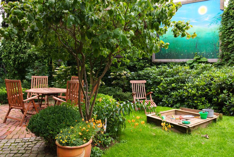 Familiengarten mit Sitzplatz unter einem Baum, üppiger Bepflanzung und Sandkasten